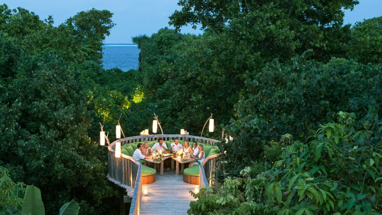 A group of people dining on a tree-top platform surrounded by lush greenery at Soneva Fushi in the Maldives. The circular, elevated deck with a wooden pathway features ambient lighting and offers a scenic view, with the ocean visible in the background.