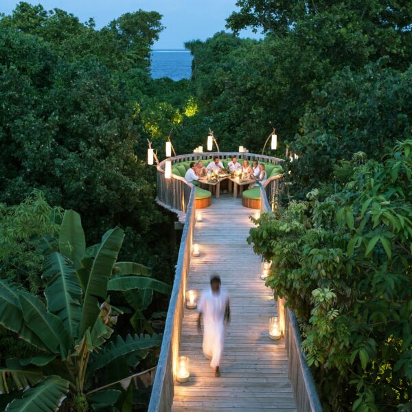 A person in white walks along a wooden pathway illuminated by candlelight amidst dense greenery, leading to a circular dining area at Soneva Fushi. Elevated above the trees with people seated under hanging lights, this Maldives dining experience offers a stunning ocean view in the background.