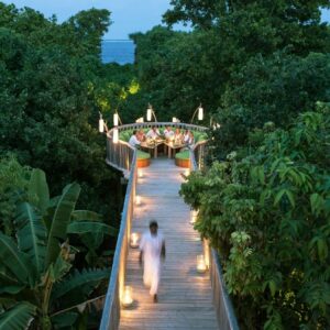 A person dressed in white walks along a wooden pathway surrounded by lush greenery at the Maldives luxury resort, Soneva Fushi. The pathway leads to a circular dining area elevated above the trees, where people are seated. Lanterns line the pathway, creating a warm, inviting ambiance during the evening at the villas.