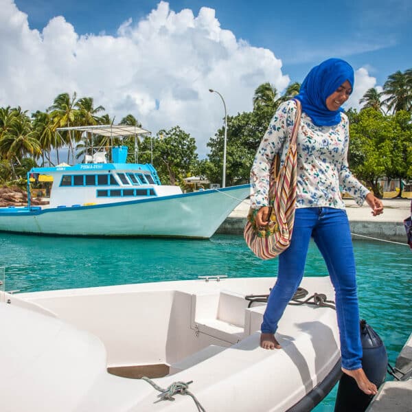 A woman in a blue hijab and jeans steps off a small boat onto a concrete dock under a partly cloudy sky, embodying the Soneva Exclusive Experience Maldives. She carries a colorful striped bag, with another person nearby carrying a backpack. Blue water and palm trees frame the bigger white and blue boat in the background.