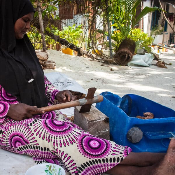 A woman in a bright, patterned dress and black headscarf sits on the ground using a wooden tool to manually process a coconut. She is surrounded by tropical foliage and small structures in the background. A large blue container is beside her, collecting the coconut meat—truly a Soneva Exclusive Experience Maldives moment.