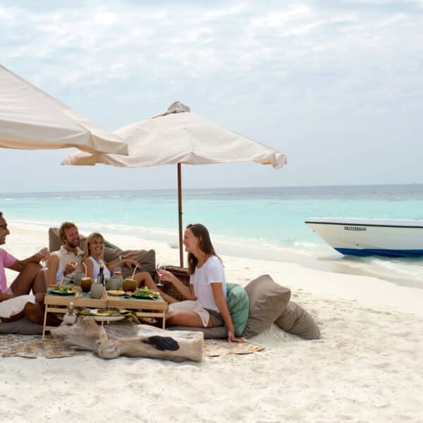 Four people sit on cushions under umbrellas, savoring a meal on a pristine beach. A table is set with food and drinks. In the background, the ocean is calm and a small boat is anchored near the shore. The sky is partly cloudy, enhancing this Maldives exclusive experience at Soneva with serene beauty.