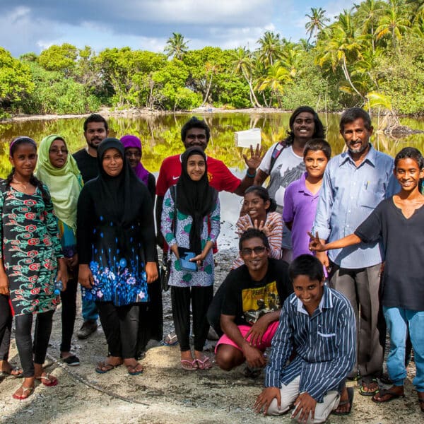 A group of 19 people, including men, women, and children, stand together outdoors with a tropical forest and a body of water in the background. They are smiling and dressed in colorful, casual attire. The sky is partly cloudy, capturing the joyful essence of a Soneva Exclusive Experience Maldives.