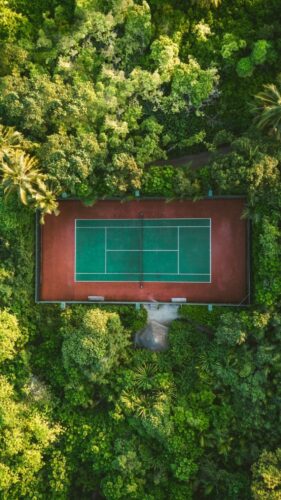 Aerial view of a tennis court with green and blue surfaces, amid dense, lush jungle foliage. Tall trees and various greenery surround the rectangular court at Soneva, creating a striking contrast between the structured sports area and the wild natural environment for an exclusive Maldives experience.