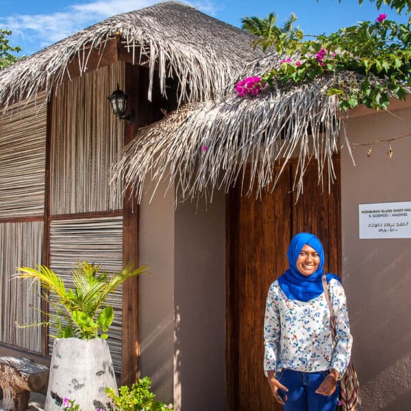 A woman in a blue hijab stands smiling in front of a tropical building with a thatched roof and wooden doors. The building, part of the Maldives Exclusive Experience by Soneva, is surrounded by lush greenery and blooming flowers. The clear blue sky suggests a sunny day.