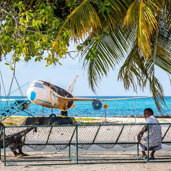 A beach scene featuring people lounging on swings under palm trees. In the background, there is an old airplane fuselage placed on the sand with the ocean in the distance. The vibrant blue water contrasts with the green leaves and the white sand, epitomizing a Maldives exclusive experience.