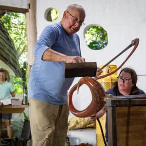 A man holding a black tool shapes molten glass as a woman assists by holding the glass item, with several onlookers in the background. They are in a workshop setting, surrounded by glassblowing equipment and tools. This Maldives Exclusive Experience at Soneva is well-lit with natural light.