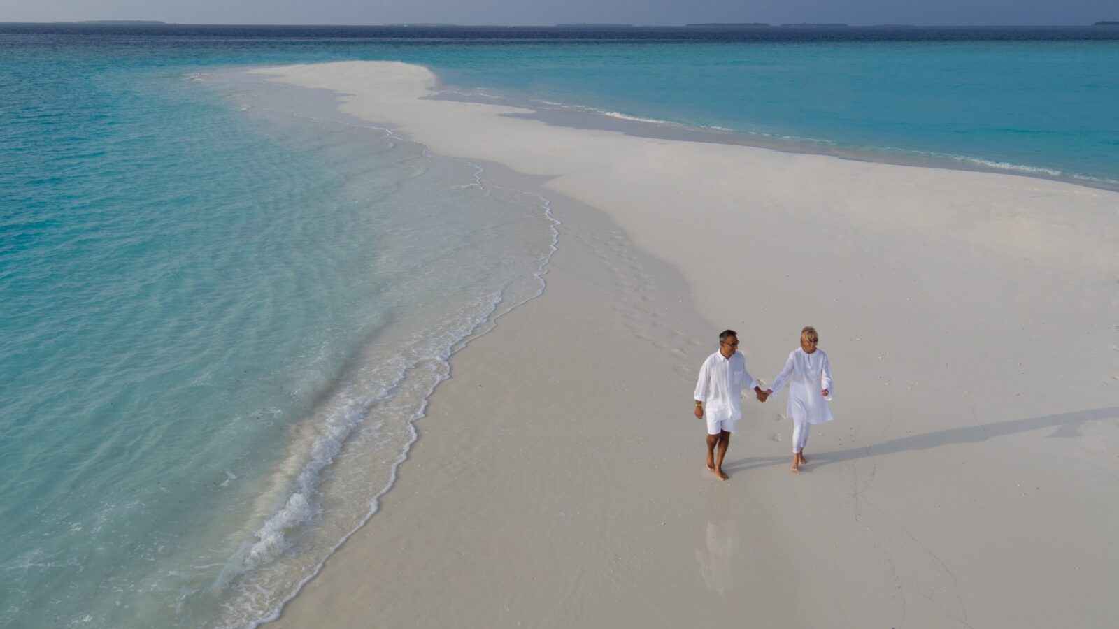 A couple walks hand-in-hand along a pristine, curved white sand beach with vibrant turquoise water on both sides at a Maldives Honeymoon Resort. They are dressed in light clothing, enjoying the serenity of the secluded tropical location under a clear sky.