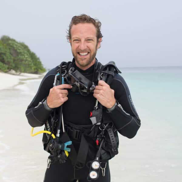 A smiling man in scuba diving gear stands on a sandy beach in the Maldives with clear blue waters and lush greenery in the background. He holds his diving mask in both hands, dressed in a black wetsuit. The sky appears clear, suggesting a pleasant day for this exclusive Soneva diving experience.