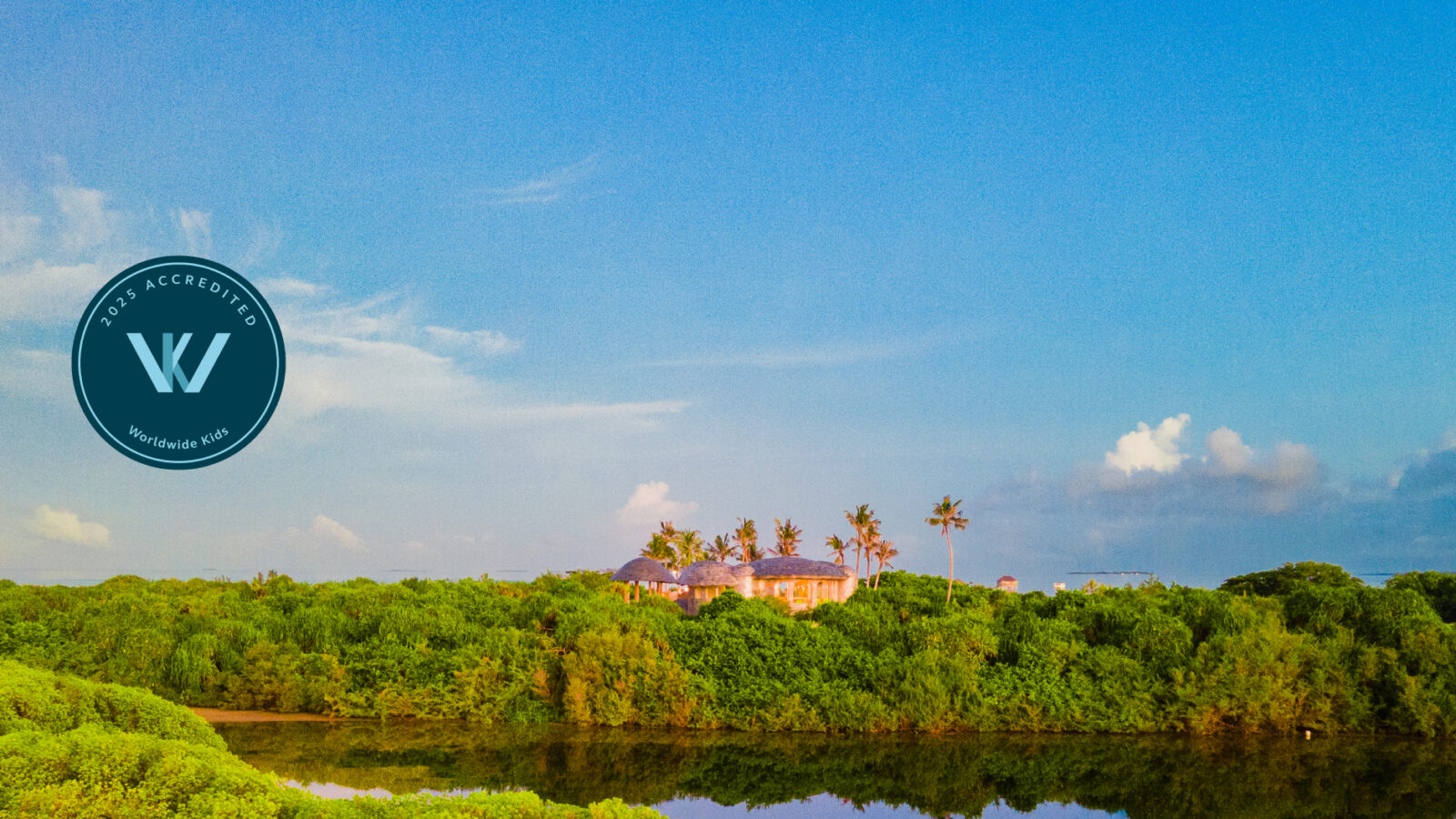 A scenic view of lush greenery with a building surrounded by trees in the distance evokes the serene charm of Soneva Jani. The sky is bright blue with scattered clouds, and a circular logo with the letter "W" graces the left side of this inviting family-friendly image.