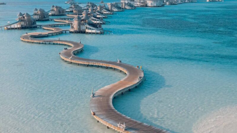 An aerial view of a long, winding wooden boardwalk curving through clear, shallow blue waters, connecting a series of overwater bungalows. The sky is mostly clear with a few clouds, and the scene gives a tranquil, tropical vibe—perfect for an unforgettable Soneva Maldives holiday.