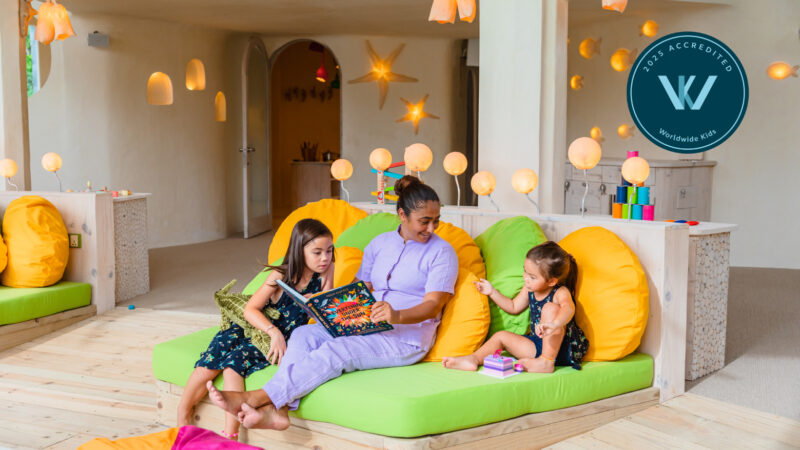 A woman sits on a lime green couch with two young girls, one with a book and the other a toy. In this vibrant Soneva Jani family room, colorful lights and toys surround them. A "W Accredited Worldwide Kids" badge is proudly displayed.