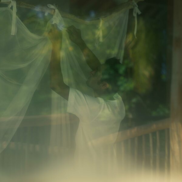 A person in a white shirt is seen through a window adjusting a sheer white curtain or mosquito net. The scene, reminiscent of the serene ambiance deserving of a Soneva Award, is partially obscured by reflections and a greenish tint, blending indoor and outdoor elements.