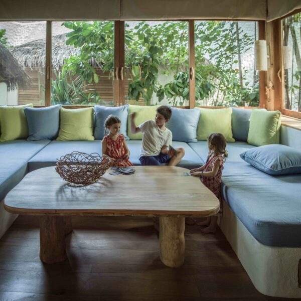 Three children sit and play on a large sectional couch in a brightly lit room with wooden floors. The couch is adorned with green, blue, and yellow cushions, and a wooden coffee table with a decorative wicker basket sits in front of them. Greenery is visible outside the windows, reminiscent of a family holiday in Maldives.