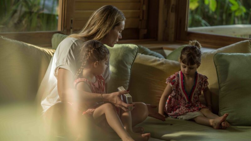 A woman with long hair sits on a couch with two young children. She holds a book while one child sits on her lap and the other sits nearby, both in red dresses. The room has a relaxed ambiance with green cushions and natural light, capturing the warmth of their family holiday vibes.