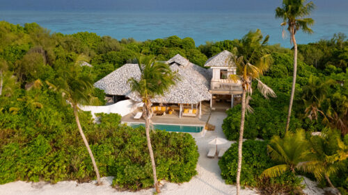 Aerial view of a luxurious beachfront hideaway surrounded by lush green palm trees. This beach villa features wooden architecture with thatched roofs, a private pool, and a sandy path leading to a pristine turquoise ocean under a clear blue sky.