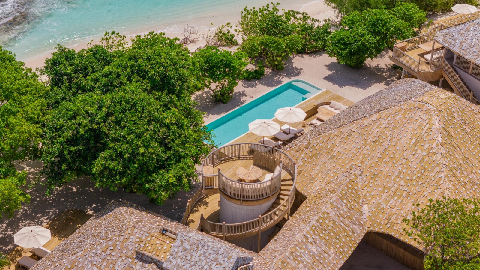 Aerial view of a tropical resort within a pristine beach reserve, featuring a curved wooden structure and spiral staircase, embraced by lush greenery. Overlooking the turquoise ocean and sandy beach, a rectangular pool with umbrellas complements the serene landscape.