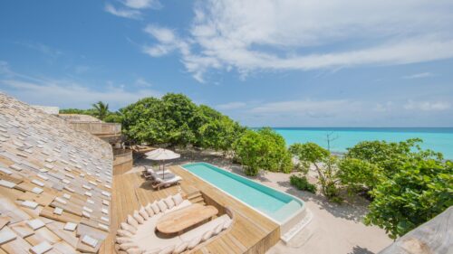 A luxurious beachfront patio at the Beach Reserve features a wooden deck surrounding an infinity pool. There's a cozy seating area with a sun umbrella. Lush greenery abounds, while the turquoise ocean and blue sky provide a serene backdrop.