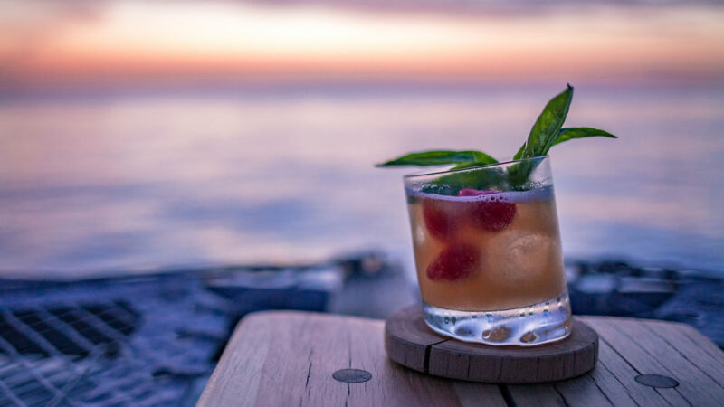 A refreshing cocktail garnished with mint leaves and raspberries sits on a wooden coaster on a table outdoors at Soneva Fushi. The background displays a serene sunset over calm water, with hues of pink, purple, and orange blending in the sky.