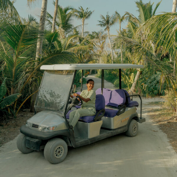 A person is driving a golf cart with a canopy roof down a dirt path surrounded by lush, tropical greenery and tall palm trees. The golf cart has purple seats, and the driver, wearing a light-colored shirt and looking towards the camera, seems to be enjoying their journey as part of their Soneva career.