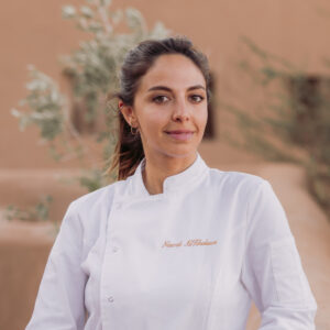 A woman with long brown hair wearing a white chef&#039;s jacket stands outdoors. The background features trees and a blurred building.