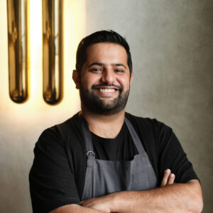 A smiling person with short dark hair and a beard stands against a textured wall, wearing a black shirt and dark-colored apron. Their arms are crossed. Two vertical light fixtures are visible on the wall to their left.