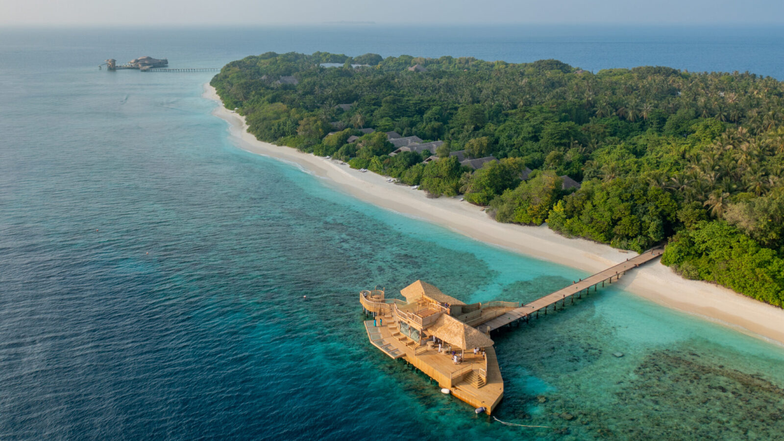 Aerial view of a tropical island featuring lush greenery, white sandy beaches, and clear turquoise waters. A wooden overwater bungalow connected by a pier extends into the ocean, surrounded by vibrant coral reefs. Another small building is seen in the distance—welcome to the Maldives Luxury Resort at Soneva Fushi Villas.