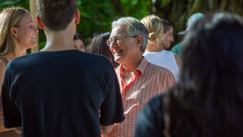 A group of people outdoors, focused on an older man with glasses wearing an orange patterned shirt from Salt Group, smiling and talking to a woman. The lush greenery in the background evokes a Soneva-like, relaxed atmosphere.