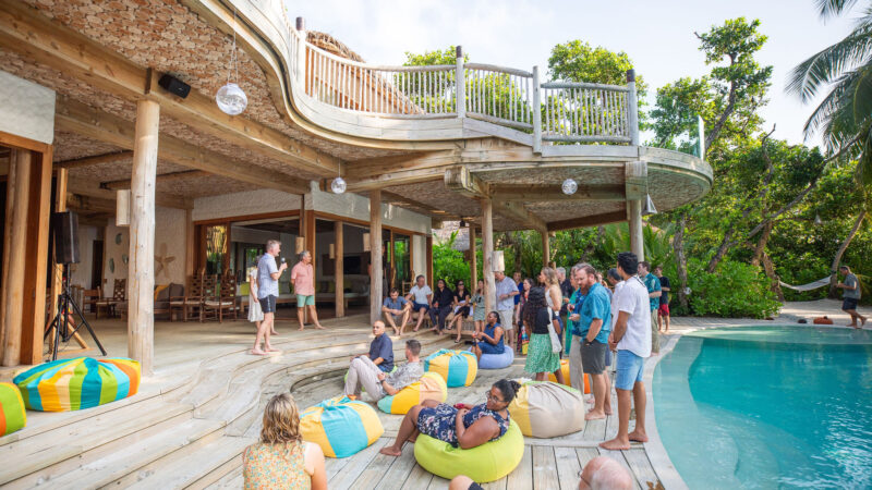 A group of people from the Salt Group gather on a wooden deck beside a curved building, surrounded by tropical plants. Some sit on colorful bean bags near a pool at Soneva. The setting is sunny, with a relaxed atmosphere.