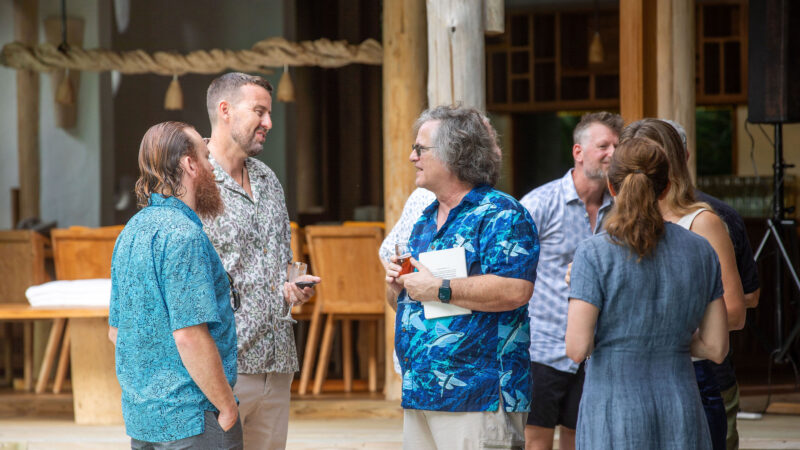 A group of people, casually dressed in patterned shirts and summer attire, are gathered outside a wooden Soneva building. Engaging in conversation, some hold papers or small devices. The relaxed setting reflects the informal vibe of a typical Salt Group gathering.