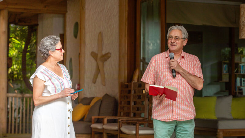 A man and woman from the Salt Group are speaking to an audience at an indoor event. The man, dressed in a pink shirt and shorts, holds a microphone and red book. The woman in a white dress holds a blue card. Comfortable seating, reminiscent of Soneva's luxurious style, is visible in the background.