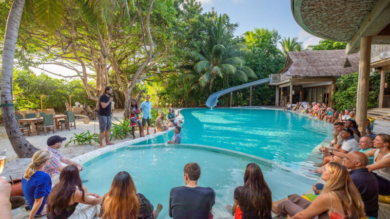 A group of people gathered around a large, curved pool at a Soneva resort, surrounded by tropical trees and buildings. Some sit at the pool's edge, others stand or swim. A waterslide is visible in the background, adding to the relaxed, tropical atmosphere curated by Salt Group designers.