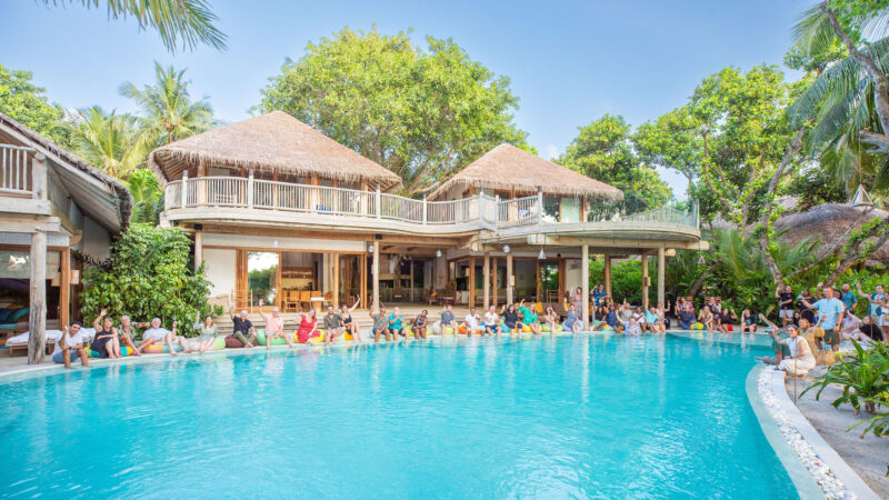 Soneva's luxurious tropical resort boasts a large blue swimming pool surrounded by lounge chairs and guests. The multi-story building features thatched roofs and open terraces. Lush greenery and palm trees from the Salt Group frame the scene under a clear blue sky.