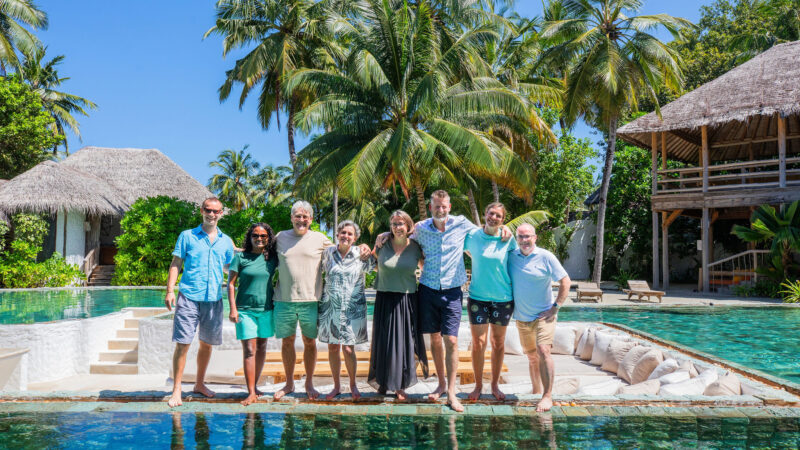 A joyful group of nine stands together, grinning by the pool in a tropical setting at Soneva. Encircled by swaying palm trees and charming thatched-roof buildings, they bask under a clear blue sky. The atmosphere is sunny and cheerful, perfectly capturing the Salt Group's adventurous spirit.