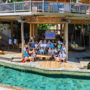 A large group of people from the Salt Group gathered on a wooden deck near a pool, in front of a rustic, multi-level building. Some sit on benches and chairs, while others stand. The setting is tropical, with lush greenery surrounding the Soneva-inspired area.