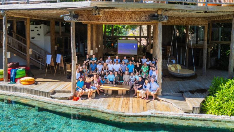 A large group of people from the Salt Group gathered on a wooden deck near a pool, in front of a rustic, multi-level building. Some sit on benches and chairs, while others stand. The setting is tropical, with lush greenery surrounding the Soneva-inspired area.