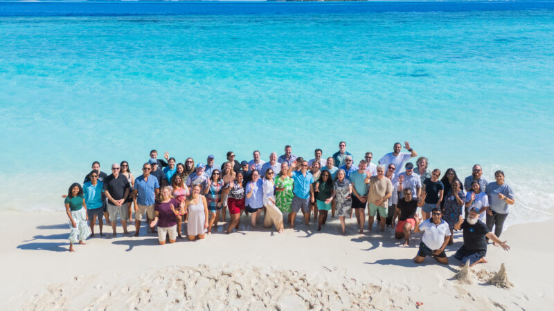 A large group of people from the Salt Group stands together on a sandy beach with Soneva's clear turquoise water in the background. Everyone is smiling and dressed in casual, summery clothing. The sky is clear with a distant island visible on the horizon.