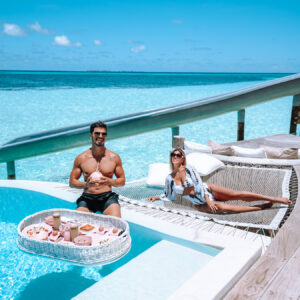 A man stands in a pool holding a floating tray of food, including drinks and pastries, while a woman lounges on a hammock beside the pool. They are both wearing swimwear, with a bright blue ocean and sky in the background, enjoying their Soneva Exclusive Offers.