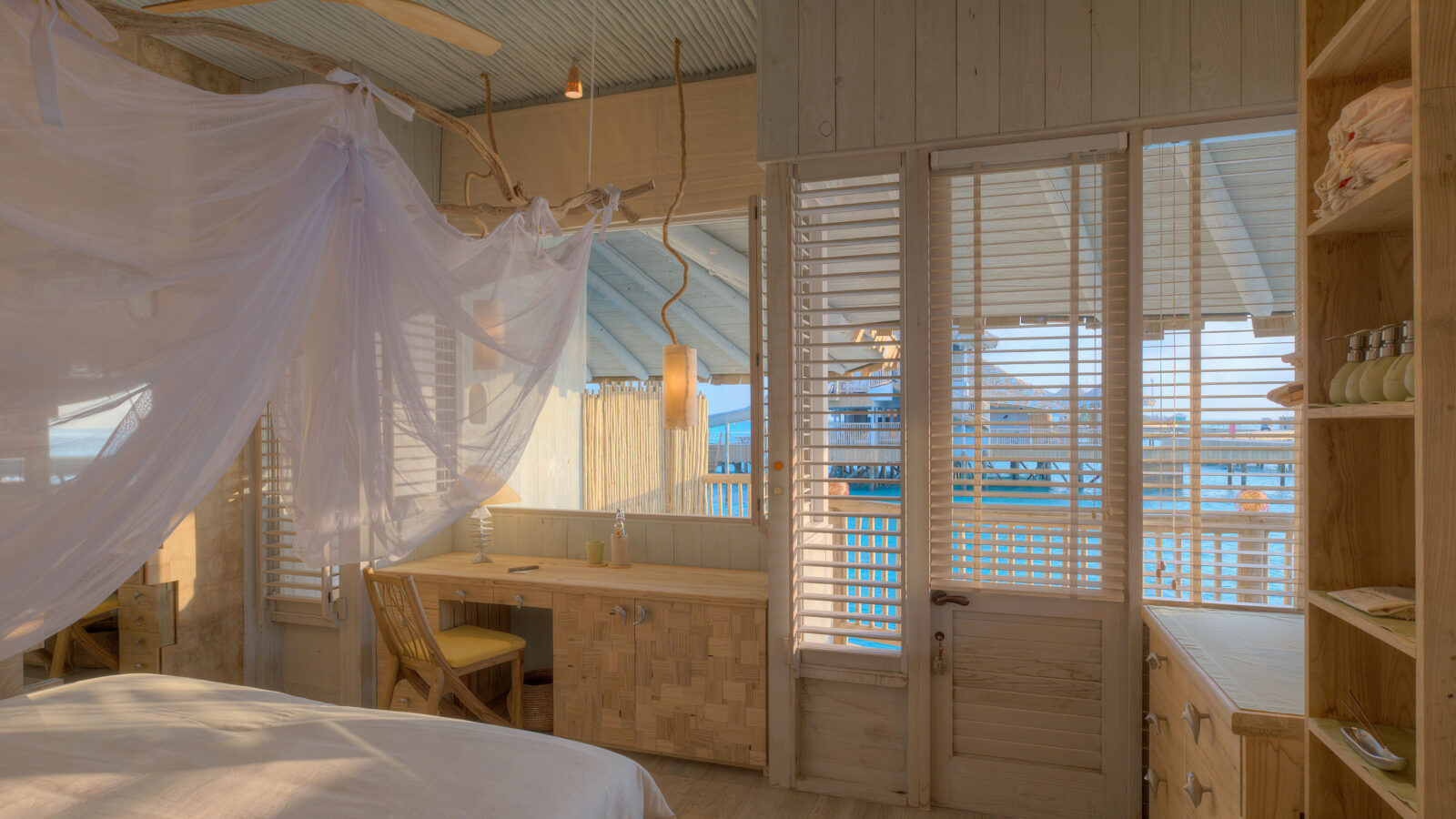 A serene, sunlit bedroom at the Crusoe Reserve features wooden decor overlooking the ocean. The room boasts a canopy bed, a desk with a chair, and large windows with shutters offering breathtaking views of the blue sea and sky outside.