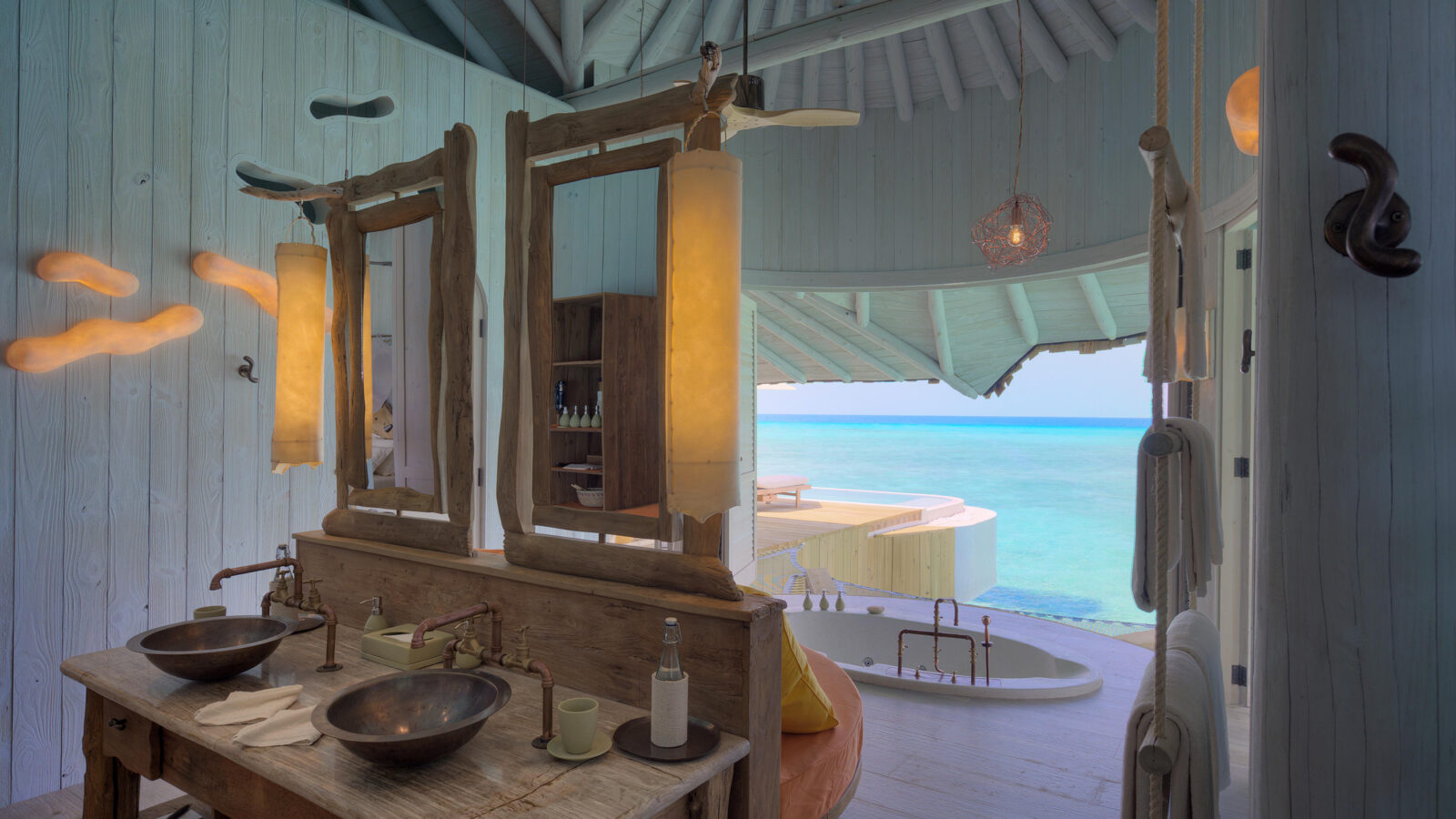 A luxurious bathroom in the Crusoe Reserve features twin wooden sinks, rustic mirrors, and a view of a spacious terrace leading to the ocean. Elegant lighting creates a warm ambiance, complemented by the turquoise sea visible through large doors.