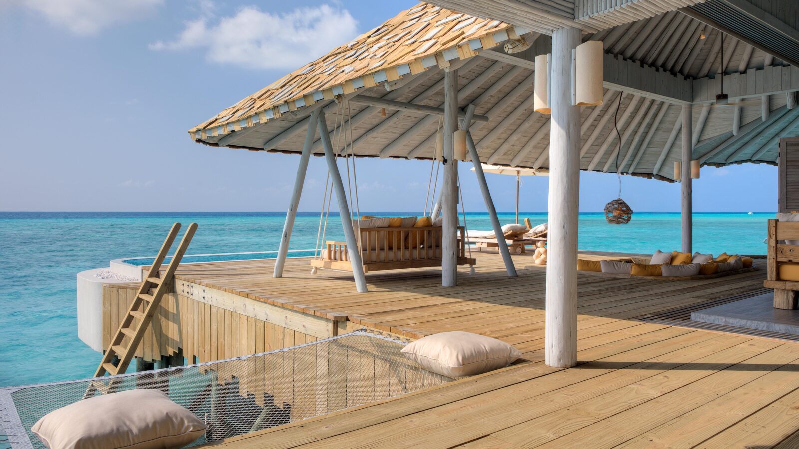 A luxurious Crusoe Reserve overwater bungalow with a thatched roof in a tropical setting, featuring wooden decking and lounge seating. It overlooks a pristine, bright blue ocean under a clear sky, with ladders leading down to the water.