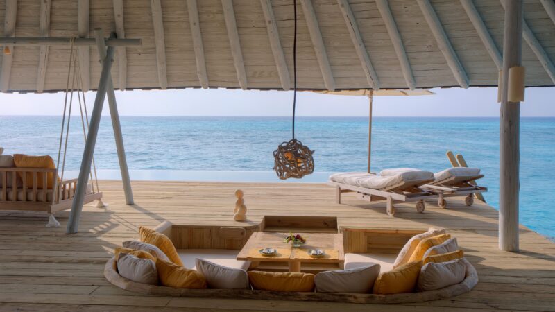 A serene seaside deck at Crusoe Reserve features sun loungers, a small pool, and a cozy seating area with cushions and a low table. A unique pendant light hangs above, while the endless blue ocean stretches out in the background.