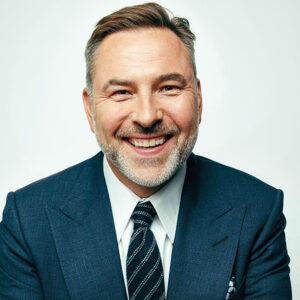 Smiling man with short hair and a beard, wearing a dark blue suit, white shirt, and dark striped tie, against a light background. His hands are crossed in front of him.