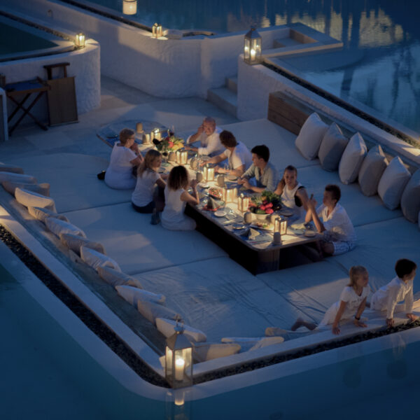 A group of people, including children and adults, are seated around a low table in a luxurious outdoor lounge area surrounded by a pool at Soneva Fushi. The Maldives evening is illuminated by numerous candles, creating a cozy and intimate atmosphere.