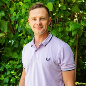 A person with short light brown hair and a purple polo shirt stands in front of a lush, green leafy background reminiscent of a Maldives luxury resort. The individual is smiling and has a relaxed demeanor.