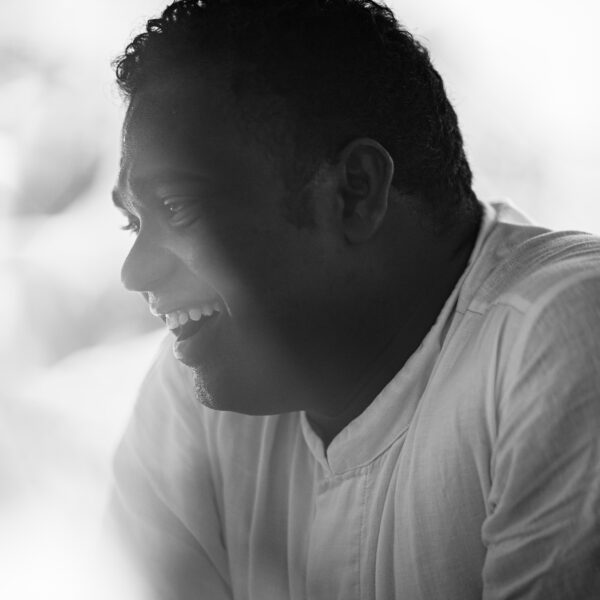 Black and white photo of a person with curly hair smiling and looking to the side. They are wearing a light-colored shirt, and the background is blurred, contributing to a serene atmosphere. The image, reminiscent of Soneva Career moments, captures a candid moment of joy.