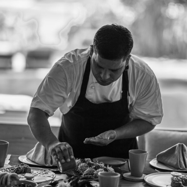 A chef in a white shirt and dark apron is meticulously plating food on a dining table in a softly lit room. The table is set with several dishes, plates, cups, and folded napkins. The background is blurred, drawing focus to the chef's skillful precision—perhaps an aspirant for a Soneva Career.