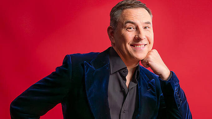 A smiling person wearing a dark velvet jacket and a button-up shirt is posing against a vibrant red background, resting their chin on their hand.