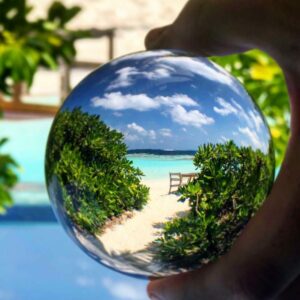 A clear spherical lens is held up against a beach backdrop, showing a magnified, inverted view of a sandy pathway leading to a turquoise sea with lush greenery on either side and a chair at the end. The scene evokes memories of a serene family holiday in Maldives' Soneva resort, surrounded by more greenery and a bright sky.