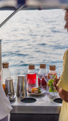 A person in a yellow shirt stands on a boat near a counter with various drinks and garnishes, including glass containers with red liquid, glassware, passion fruits, and herbs. The ocean of the Maldives is visible in the background, capturing an exclusive Soneva experience.